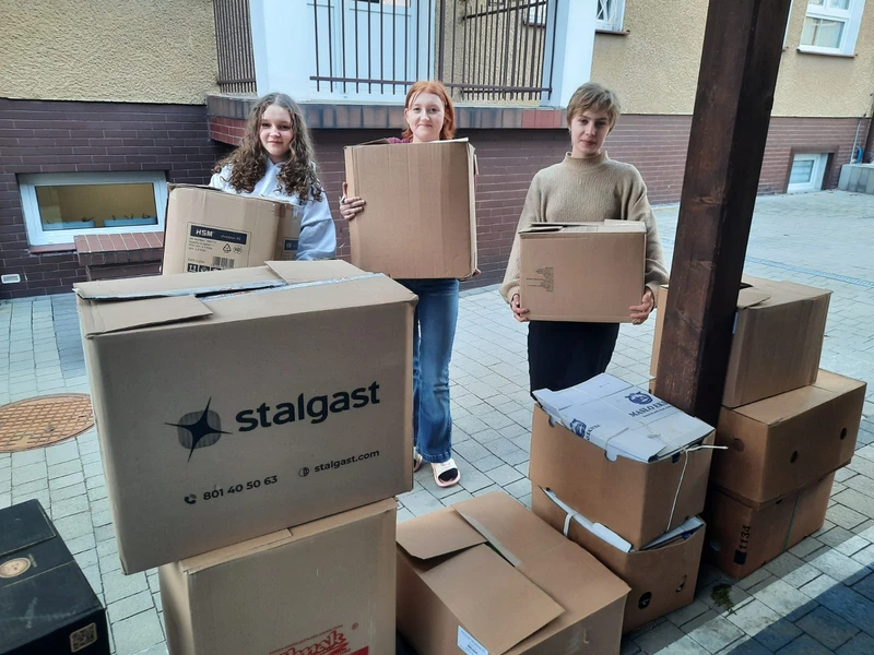 Na zdjęciu widać trzy młode osoby stojące na chodniku przed budynkiem mieszkalnym. Każda z nich trzyma w rękach duże kartonowe pudełko, jakby przenosiły lub pomagały w przeprowadzce czy zbiórce rzeczy.