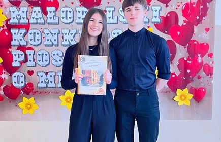 na zdjęciu widać dwie osoby chłopca i dziewczynę w pozycji stojącej obok siebie ,dziewczyna trzyma w rękach dyplom .Obydwoje stoją przed ścianą na kt&oacute;rej wisi plakat z napisem XII Małopolski Konkurs Piosenki o Miłości