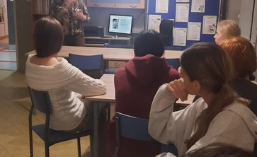  Grupa osób uczestniczących w spotkaniu lub zajęciach w sali edukacyjnej. Kilka osób siedzi przy stołach, zwróconych w stronę prowadzącej, która stoi z przodu pomieszczenia. Kobieta prowadząca prezentację gestykuluje ręką i mówi do uczestników.