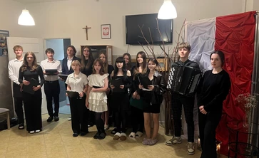 Na zdjęciu widać grupę młodych osób stojących razem w pomieszczeniu, prawdopodobnie w szkole lub domu kultury. Większość z nich ubrana jest elegancko – w ciemne lub biało-czarne stroje, co może sugerować udział w jakimś występie, akademii lub uroczystości. Jedna z osób trzyma akordeon, co wskazuje, że częścią wydarzenia mogła być muzyka.

W tle widoczna jest flaga w biało-czerwonych barwach oraz dekoracje przypominające gałązki drzew, co może sugerować temat patriotyczny lub świąteczny. Na ścian