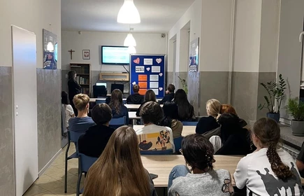 grupa uczniów siedzących przy kilku stołach w  sali, która jest jasna, schludna i dobrze oświetlona. Na ścianie po prawej stronie stoi duży niebieski baner z białym napisem: „OBRONNOŚĆ – BEZPIECZEŃSTWO”.