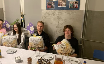 Na zdjęciu widać cztery osoby siedzące przy stole w sali, która wygląda jak szkolna lub świetlicowa. Każda z nich trzyma zapakowany prezent z napisem „Idź odważnie przez swoje życie”. Na stole stoją talerzyki, kubki, ciasto, sok i przekąski. W tle są dekoracje z wielkanocnymi jajkami oraz tablica ze zdjęciami. Atmosfera wygląda na uroczystą i miłą