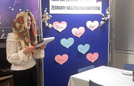 Na zdjęciu widać długi st&oacute;ł nakryty do poczęstunku . Obok stołu  jedna osoba stoi i czyta . Obok jest tablica z napisem o pożegnaniu maturzyst&oacute;w.