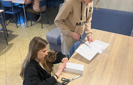 Na zdjęciu widać dwie młode dziewczyny  przy drewnianym stole w pomieszczeniu przypominającym salę lub korytarz. Jedna z nich ma na sobie jasną bluzę i jest pochylona nad kartkami, druga ubrana jest na ciemno i r&oacute;wnież skupia się na dokumentach leżących na stole.