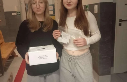 dwie uczennice stojące w korytarzu z urną do głosowania