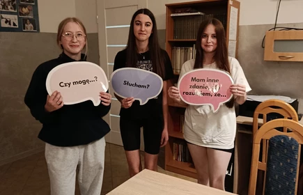Na zdjęciu widać trzy młode osoby stojące w pomieszczeniu, prawdopodobnie w sali szkolnej lub świetlicy. Każda z nich trzyma w rękach tabliczkę w kształcie dymku z napisem:

pierwsza osoba z lewej ma napis „Cześć!”,
środkowa osoba — „Dziękuję!”,
osoba po prawej — „Proszę!”.