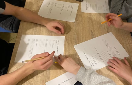 na zdjęciu widac blat drewniany na ktorym oparte są ręce uczni&oacute;w a przy nich karty pracy