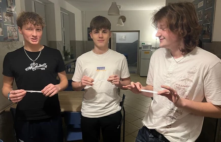 Trzech uczniów stoi obok siebie trzymając papierowe paski w rękach  