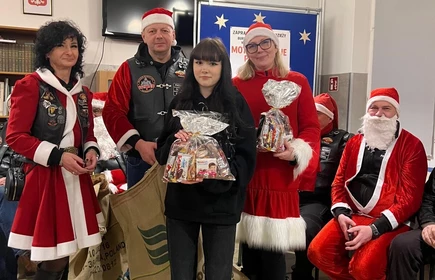 Na zdjęciu widać grupę os&oacute;b podczas świątecznego spotkania. Kilka z nich jest ubranych w stroje Mikołaja lub w czerwono-białe stylizacje. Wszyscy wyglądają na uśmiechniętych i trzymają paczki z upominkami, co sugeruje akcję charytatywną lub mikołajkową. Scena odbywa się w pomieszczeniu z dekoracjami świątecznymi.