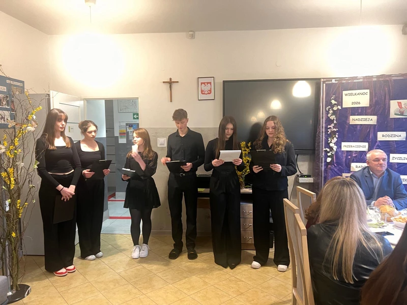 Po lewej stronie stoją  dziewczyny ubrane na czarno i i chłopiec trzymają kartki  i czytają tekst z kartek i prezentują krótką część artystyczną

Za nimi znajduje się dekoracyjna plansza w fioletowym kolorze. Widać na niej napisy: „Wielkanoc 2026”, „Nadzieja”, „Rodzina”, „Przebaczenie”, „Radość”. Ozdobiona jest też wiosennymi elementami.Po prawej stronie  widac dwie dorosłe osoby
