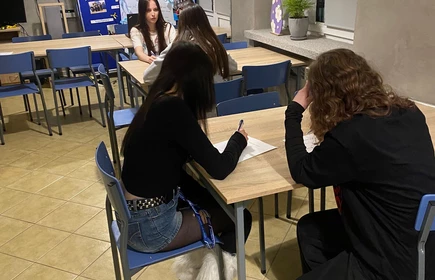 zdjęcie przedstawia  uczennice siędzace przy stolikach  in pochylajace się nad białymi kartkami uczennice w ręku trzymaja długopis