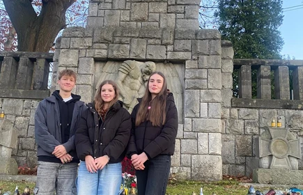 Na zdjęciu stoją trzy osoby przed kamiennym pomnikiem. Mają na sobie ciepłe ubrania, a przed nimi ustawione są znicze i kwiaty. Scena wygląda na spokojną i upamiętniającą.