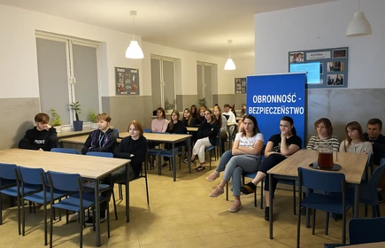 Uczniowie siedzą na niebieskich krzesłach przy prostokątnych stołach ustawionych w rzędach. Przed nimi, na końcu sali, znajduje się tablica i ekran, na którym wyświetlana jest prezentacja. Obok ekranu wisi krzyż oraz godło Polski. Na ścianie pod ekranem widoczna jest półka z książkami, a obok duża, niebieska tablica z kolorowymi kartkami i serduszkami – wygląda na planszę informacyjną lub projekt edukacyjny.
Wszyscy uczniowie zwróceni są w stronę prezentacji, słuchając osoby prowadzącej.