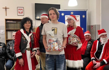 Na zdjęciu widać grupę os&oacute;b podczas świątecznego spotkania. Kilka z nich jest ubranych w stroje Mikołaja lub w czerwono-białe stylizacje. Wszyscy wyglądają na uśmiechniętych i trzymają paczki z upominkami, co sugeruje akcję charytatywną lub mikołajkową. Scena odbywa się w pomieszczeniu z dekoracjami świątecznymi.