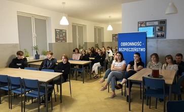 Uczniowie siedzą na niebieskich krzesłach przy prostokątnych stołach ustawionych w rzędach. Przed nimi, na końcu sali, znajduje się tablica i ekran, na którym wyświetlana jest prezentacja. Obok ekranu wisi krzyż oraz godło Polski. Na ścianie pod ekranem widoczna jest półka z książkami, a obok duża, niebieska tablica z kolorowymi kartkami i serduszkami – wygląda na planszę informacyjną lub projekt edukacyjny.
Wszyscy uczniowie zwróceni są w stronę prezentacji, słuchając osoby prowadzącej.