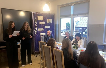 Po lewej stronie stoją dwie dziewczyny ubrane na czarno. Jedna trzyma kartkę i czyta, a druga ma w rękach teczkę i r&oacute;wnież wygląda, jakby przygotowała wystąpienie. Sprawiają wrażenie, że składają życzenia albo prezentują kr&oacute;tką część artystyczną ????????.

Za nimi znajduje się dekoracyjna plansza w fioletowym kolorze. Widać na niej napisy: &bdquo;Wielkanoc 2026&rdquo;, &bdquo;Nadzieja&rdquo;, &bdquo;Rodzina&rdquo;, &bdquo;Przebaczenie&rdquo;, &bdquo;Radość&rdquo;. Ozdobiona jest też wiosennymi elementami.Po prawej stronie sa siedzące dorosłe osoby
