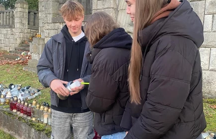 na zdjęciu stoją trzy osoby przed kamiennym pomnikiem zapalające znicz