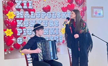 na zdjęciu widoczne są dwie osoby chłopiec siedzący na krzesle  na głowie ma kapelusz a w rekach trzyma akordeon  jest skierowany do stojącej dziewczyny która w prawej ręce trzyma mikrofon obydwoje stoją przed plakatem zawisnietym na białej scianie na którym widnieje napis XII Małopolski Konkurs Piosenki o Miłości 