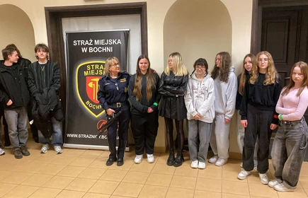 Na zdjęciu widać grupę młodzieży stojącą w pomieszczeniu, prawdopodobnie w budynku instytucji publicznej. W centrum znajduje się kobieta w mundurze straży miejskiej, która wygląda na osobę prowadzącą spotkanie lub prelekcję. Obok niej ustawiony jest stojący baner z napisem „Straż Miejska w Bochni” oraz herbem formacji.