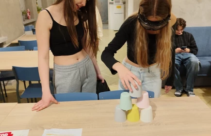 Dwie młode kobiety stoją przy stole z założonymi opaskami na oczach i pr&oacute;bują ustawiać kolorowe kubki. To wygląda na ćwiczenie grupowe lub warsztatowe w sali szkolnej.