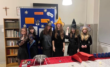 ChatGPT powiedział:

Na zdjęciu widać grupę sześciu młodych osób przebranych w stroje inspirowane andrzejkowymi wróżbami — wszystkie są ubrane na czarno, a część z nich ma na głowach ozdobne kapelusze czarownic w różnych kolorach i wzorach. Stoją obok siebie, uśmiechnięte lub pozujące, na tle dużej niebieskiej tablicy dekoracyjnej z napisem „ANDRZEJKI”, gwiazdkami oraz ilustracjami związanymi z tradycyjnymi wróżbami.