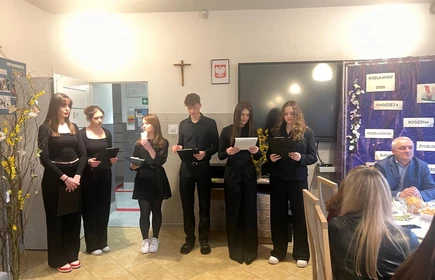 Po lewej stronie stoją  dziewczyny ubrane na czarno i i chłopiec trzymają kartki  i czytają tekst z kartek i prezentują kr&oacute;tką część artystyczną

Za nimi znajduje się dekoracyjna plansza w fioletowym kolorze. Widać na niej napisy: &bdquo;Wielkanoc 2026&rdquo;, &bdquo;Nadzieja&rdquo;, &bdquo;Rodzina&rdquo;, &bdquo;Przebaczenie&rdquo;, &bdquo;Radość&rdquo;. Ozdobiona jest też wiosennymi elementami.Po prawej stronie  widac dwie dorosłe osoby
