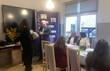 Po lewej stronie stoi młoda dziewczyna ubrana na czarno. Trzyma w rękach ozdobny kosz prezentowy z upominkiem i wręcza go starszemu mężczyźnie w garniturze. Mężczyzna uśmiecha się Po prawej stronie przy stole siedzi kilka os&oacute;b. Dwie kobiety rozmawiają ze sobą i uśmiechają się, a pozostałe osoby siedzą i obserwują sytuację. Na stole znajdują się potrawy, talerze i dekoracje, co wskazuje na wsp&oacute;lny, świąteczny poczęstunek.