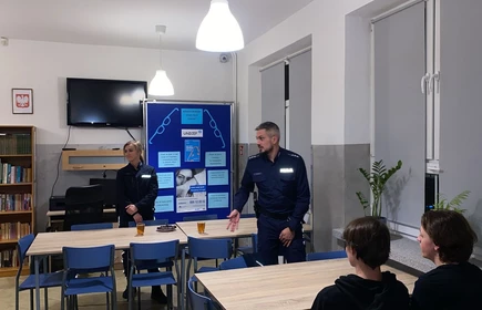 Przy kilku ustawionych obok siebie stołach siedzi jedna osoba (widoczna od tyłu), a dwie inne stoją przy stołach. Te dwie osoby mają na sobie ciemne, jednakowe stroje służbowe, co sugeruje, że mogą prowadzić zajęcia, spotkanie lub prezentację.

W tle stoi tablica ekspozycyjna z kilkoma plakatami i wydrukami, które wyglądają na materiały edukacyjne.