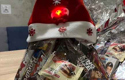 Na zdjęciu widać grupę os&oacute;b podczas świątecznego spotkania. Kilka z nich jest ubranych w stroje Mikołaja lub w czerwono-białe stylizacje. Wszyscy wyglądają na uśmiechniętych i trzymają paczki z upominkami, co sugeruje akcję charytatywną lub mikołajkową. Scena odbywa się w pomieszczeniu z dekoracjami świątecznymi.