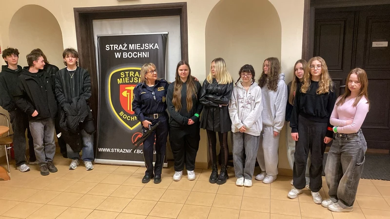 Na zdjęciu widać grupę młodzieży stojącą w pomieszczeniu, prawdopodobnie w budynku instytucji publicznej. W centrum znajduje się kobieta w mundurze straży miejskiej, która wygląda na osobę prowadzącą spotkanie lub prelekcję. Obok niej ustawiony jest stojący baner z napisem „Straż Miejska w Bochni” oraz herbem formacji.