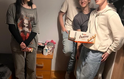 Na zdjęciu widać czworo młodych ludzi stojących w niewielkim pokoju. Trzy osoby (chłopcy) stoją razem przy ścianie po prawej stronie &ndash; jeden z nich trzyma pudełko baton&oacute;w Kinder Bueno. Wszyscy wyglądają na uśmiechniętych i rozluźnionych, jakby pozowali do zdjęcia w przyjaznej, domowej atmosferze.