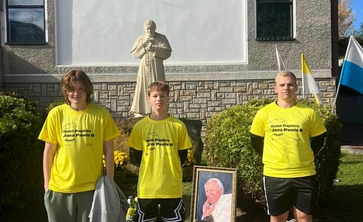 trzech uczniów w żółtych podkoszulkach  stojących przed pomnikiem Papieża