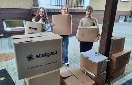 Na zdjęciu widać trzy młode osoby stojące na chodniku przed budynkiem mieszkalnym. Każda z nich trzyma w rękach duże kartonowe pudełko, jakby przenosiły lub pomagały w przeprowadzce czy zbiórce rzeczy.