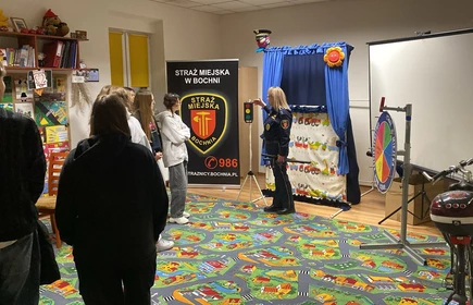 Młodzi ludzie stoją przed  funkcjonariuszką w mundurze strazy miejskiej &ndash; część z nich po lewej,stronie. Ubrani są na co dzień, gł&oacute;wnie w ciemne bluzy, spodnie dresowe i kurtki. Niekt&oacute;rzy mają ręce skrzyżowane lub trzymają się swobodnie, co sugeruje luźną, ale zorganizowaną sytuację, np. zajęcia edukacyjne lub spotkanie informacyjne.