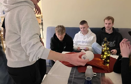 dwóch  stojących uczniów przed stołem z wróżbami obok trzech uczniów siedzących na kanapie